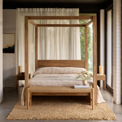 A rustic four-poster canopy bed made of natural light wood, styled in a modern minimalist bedroom with a woven bench, neutral bedding, and a jute rug.