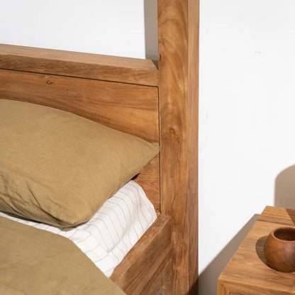 Close-up of a wooden bed frame corner with a tan pillow, white striped bedding, and part of a matching wooden bedside table against a white wall.