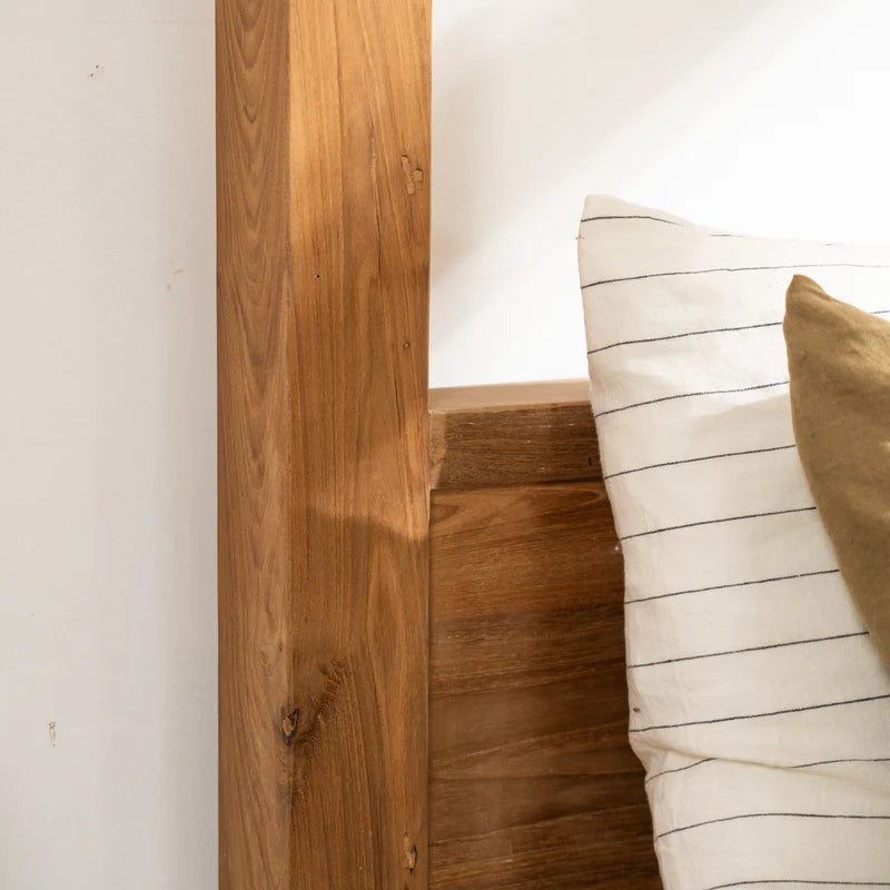 Close-up of a wooden bed frame headboard and side rail with a cream pillow featuring thin black stripes and a tan accent pillow against a white wall.