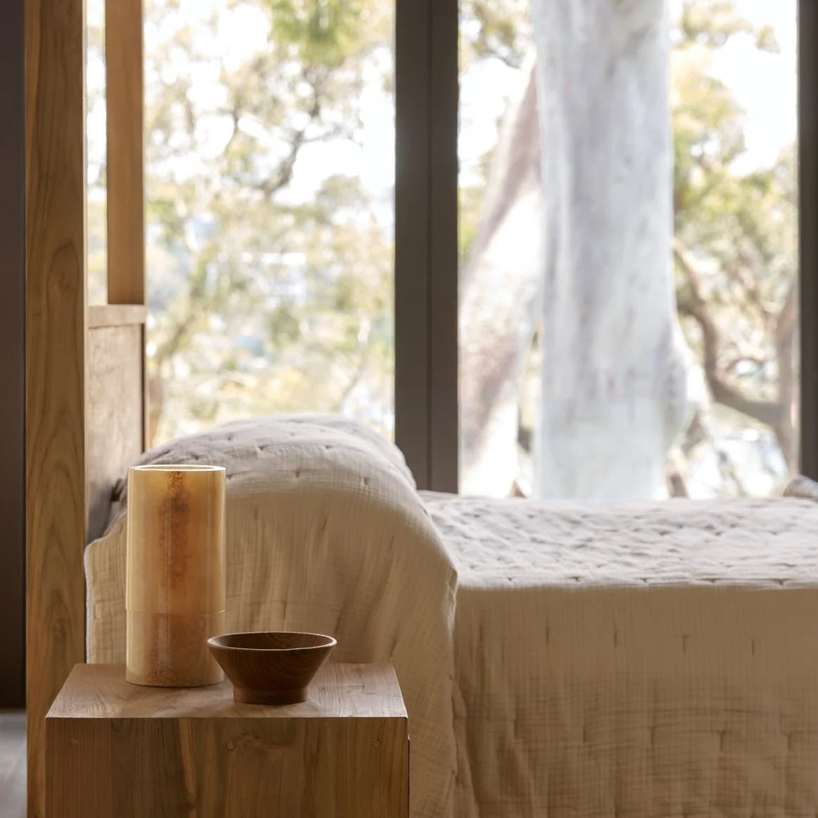 A softly lit bedroom scene with a bed dressed in textured cream bedding, a wooden side table, a tall natural wood candle, and a small dark bowl. Large windows and blurred trees are visible in the background.