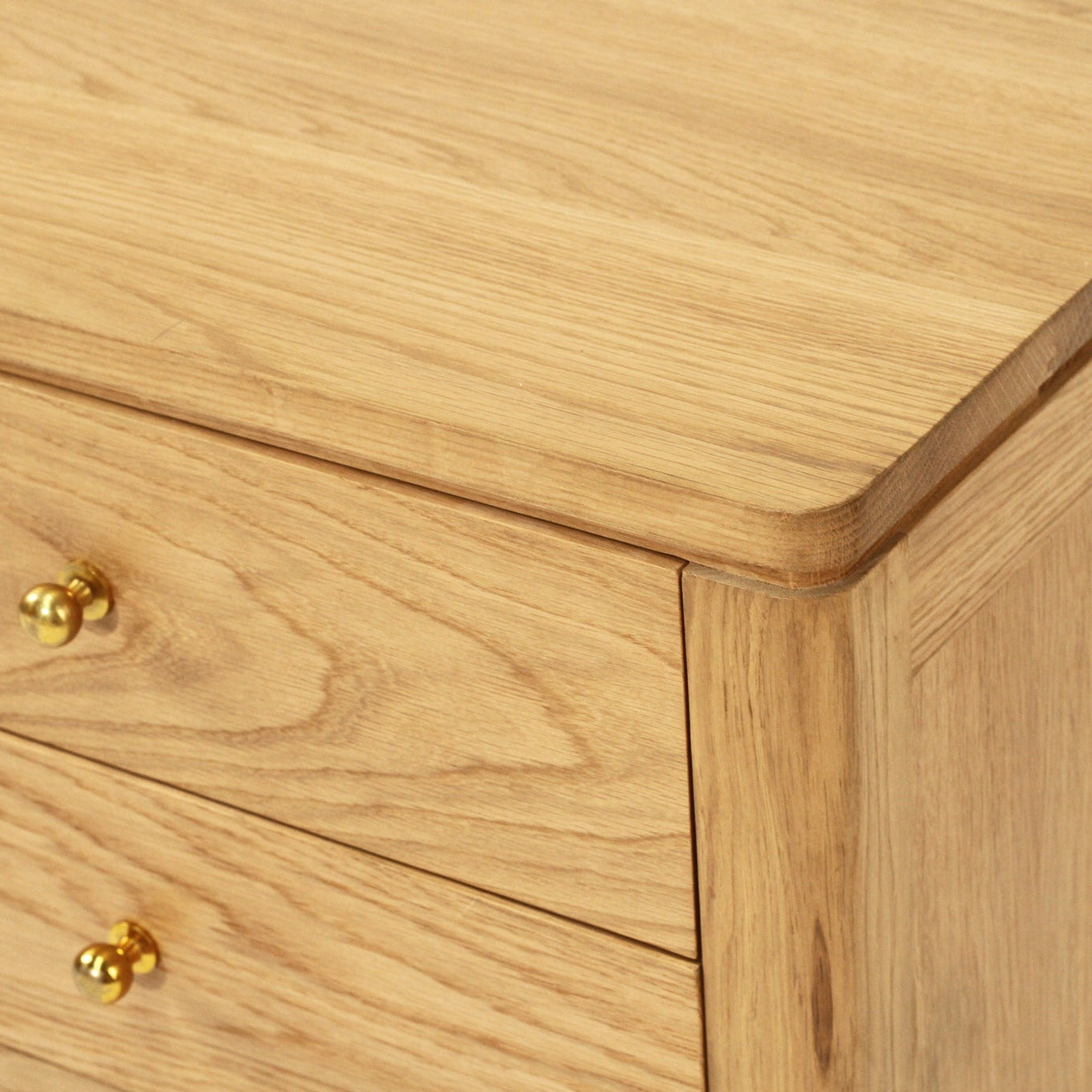 Close-up of a light oak wood cabinet or dresser showing the grain, rounded corners, and two brass drawer knobs.