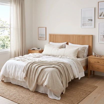 A neatly made bed in a bright, minimalist bedroom featuring a light wood slatted headboard, neutral beige and white bedding, matching bedside tables, and a woven rug.