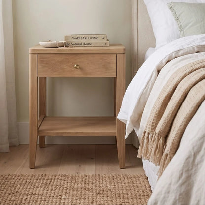A light oak wood nightstand with a single drawer and open lower shelf, styled next to a bed with white linens and a beige throw blanket in a softly lit bedroom.