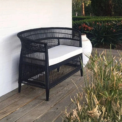 Black wicker bench with white cushion on a wooden deck with garden view