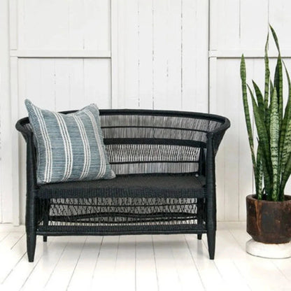 Black wicker loveseat with a striped pillow on a white wooden floor, next to a potted plant.