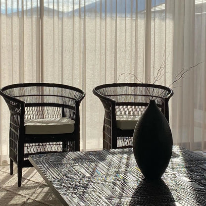 Two black Malawi Cane chairs with white cushions on a patterned rug in a sunlit room.