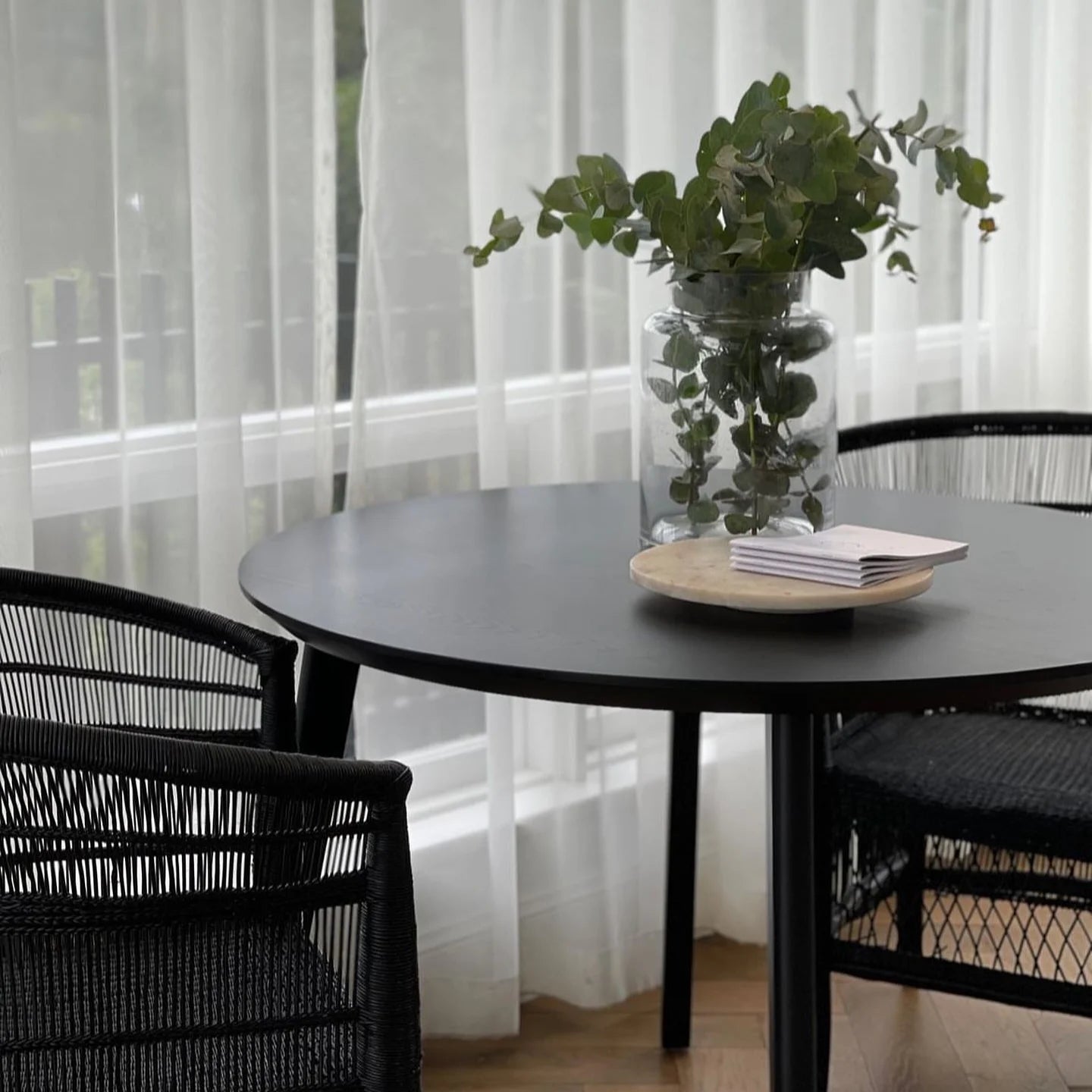 Black round table with a vase of greenery and books, surrounded by black chairs in a room with white curtains.
