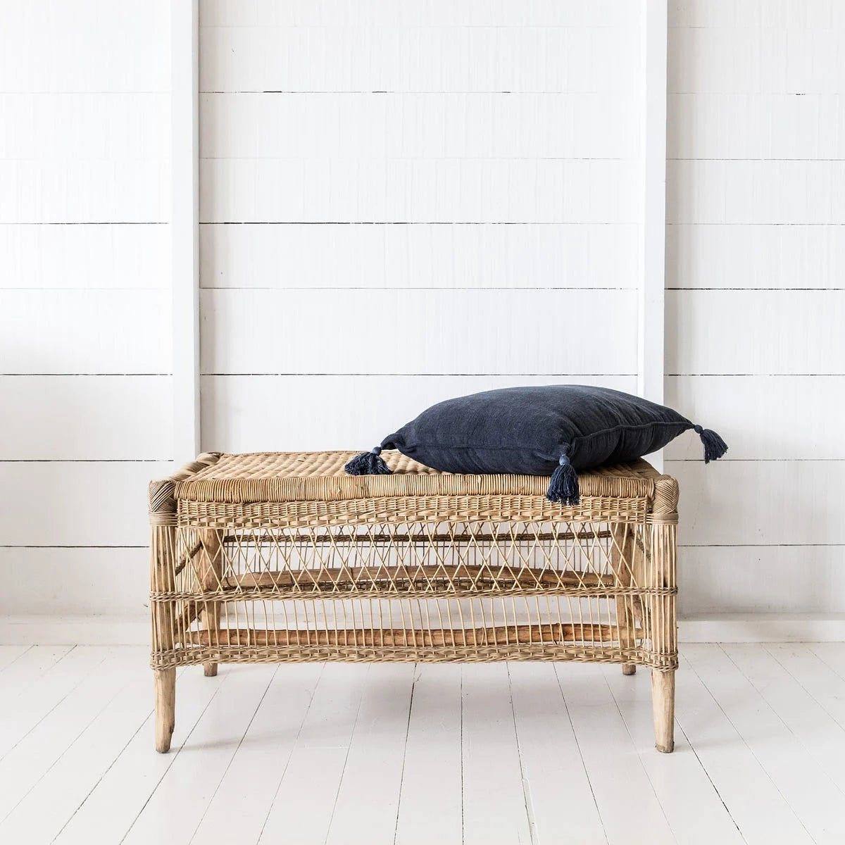 Wicker bench with a dark blue cushion against a white wooden wall.