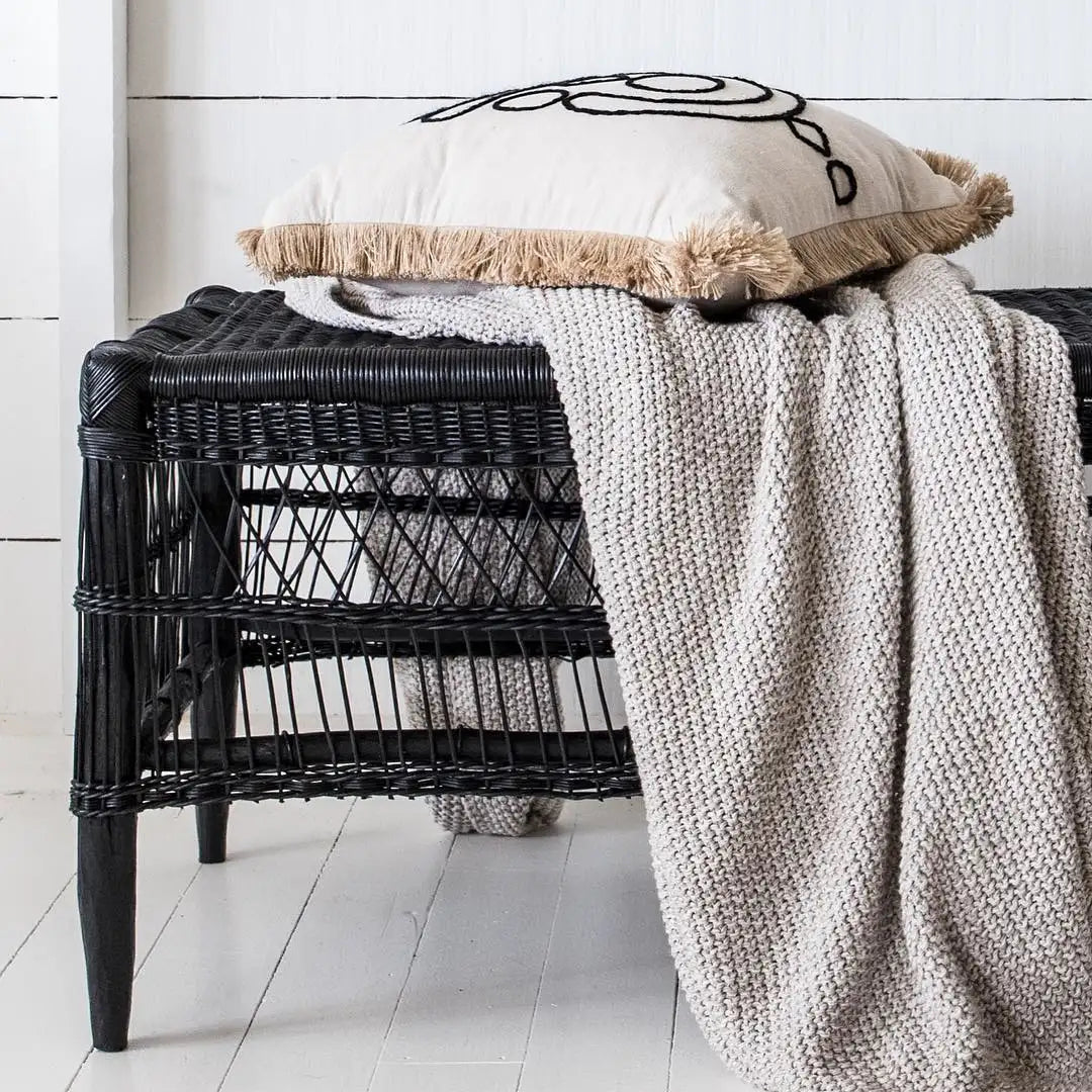 Black wicker bench with a beige cushion and knitted blanket on a white wooden floor.