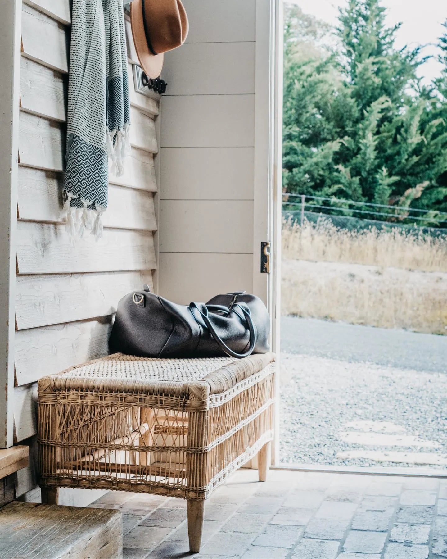 Wicker bench with a cushion and leather bag against a wooden wall with an open door leading to a garden.