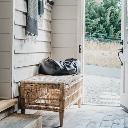 Wicker bench with a cushion and leather bag against a wooden wall with an open door leading to a garden.