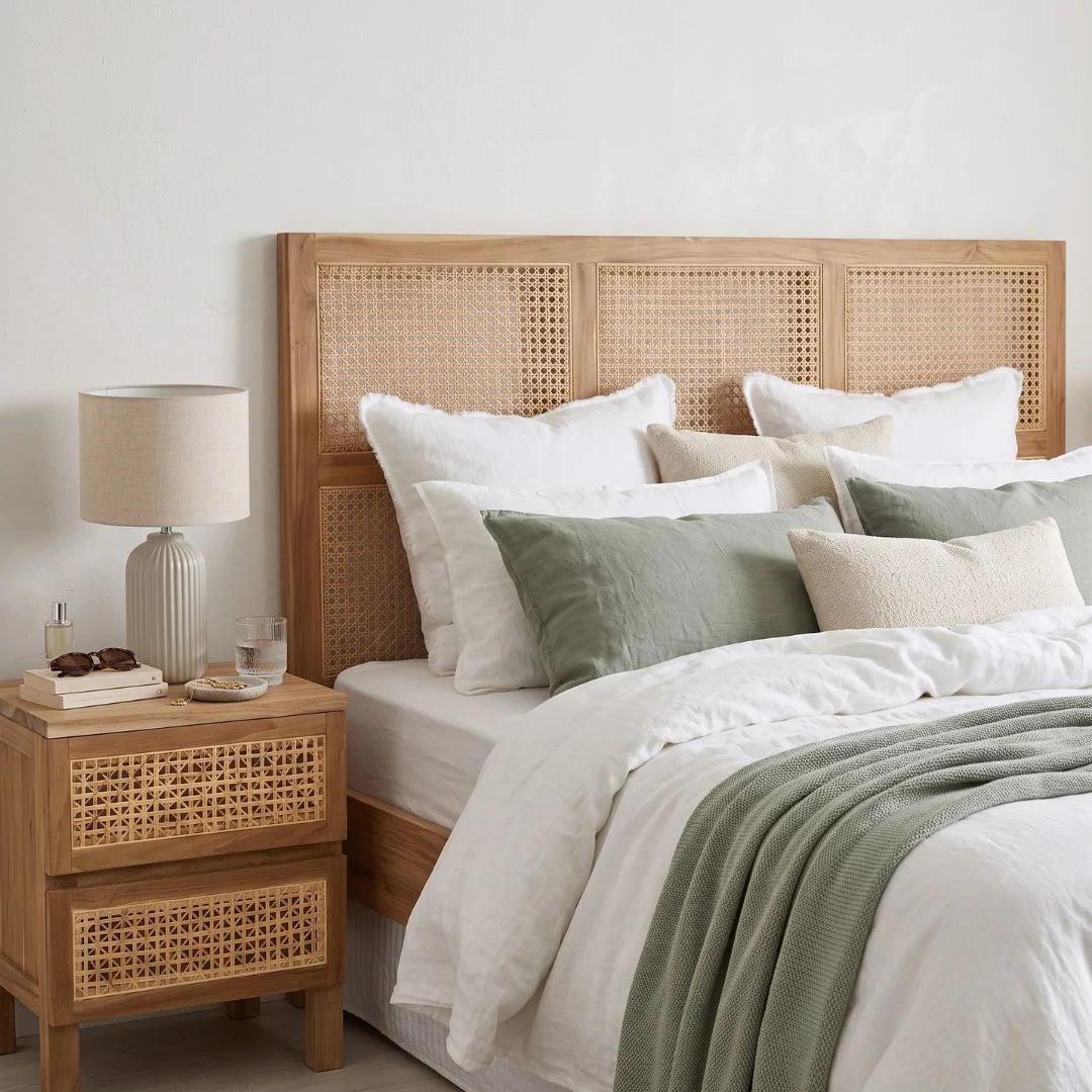 A styled bedroom scene showing a wooden bed frame with a woven cane headboard, dressed in white linens and sage green pillows and throw, next to a matching wooden nightstand with a lamp.