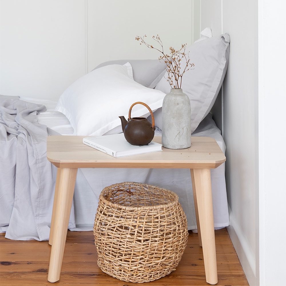 Wooden side table with a teapot, vase, and notebook next to a bed with gray bedding.