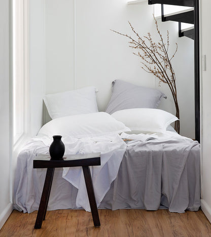 Bedroom with a bed featuring white pillows and gray bedding, a small wooden table with a black vase, and a plant against a white wall.