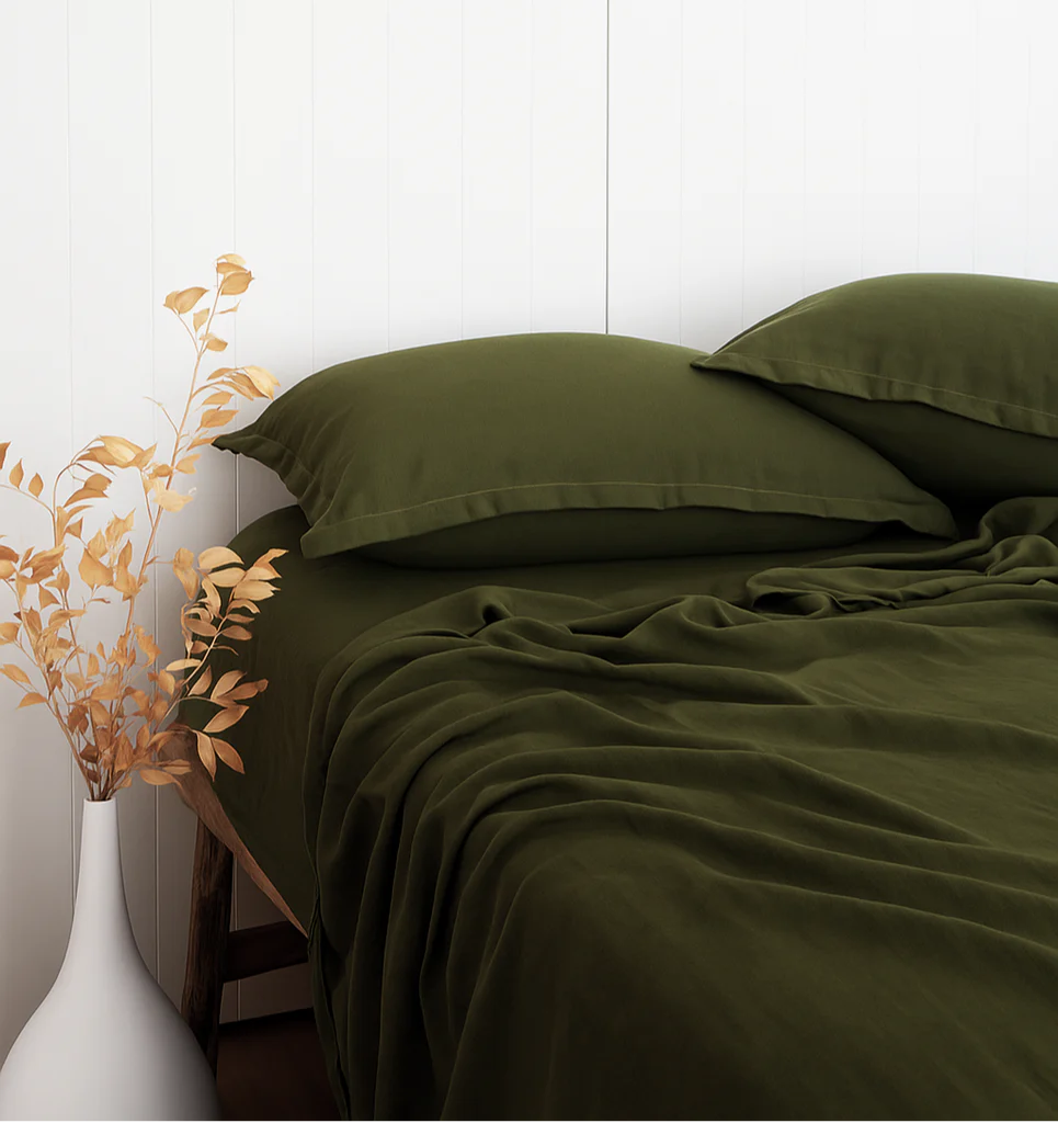 Green bed sheets and pillows on a bed with a white wall and decorative plant in the background.