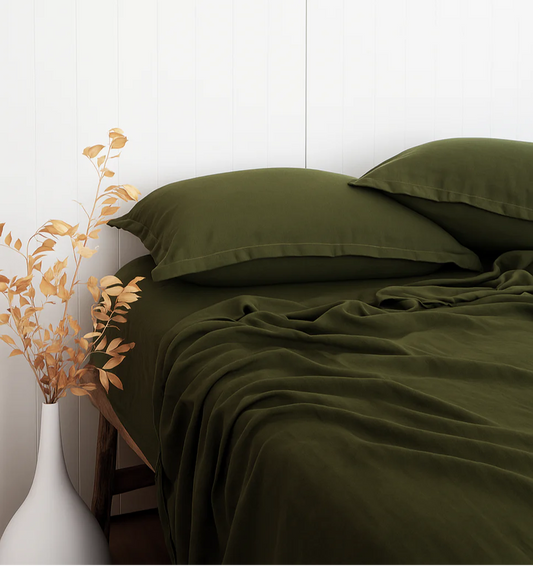 Green bed sheets and pillows on a bed with a white wall and decorative plant in the background.