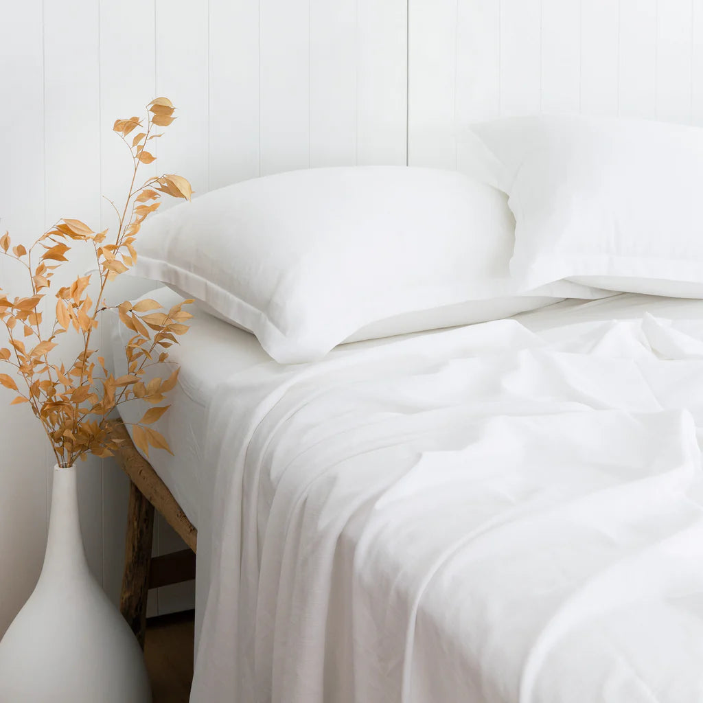 White bedding set on a bed with a vase of dried flowers on a white surface.