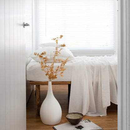 White vase with gold leaves on a wooden stool in a room with white walls and a window.
