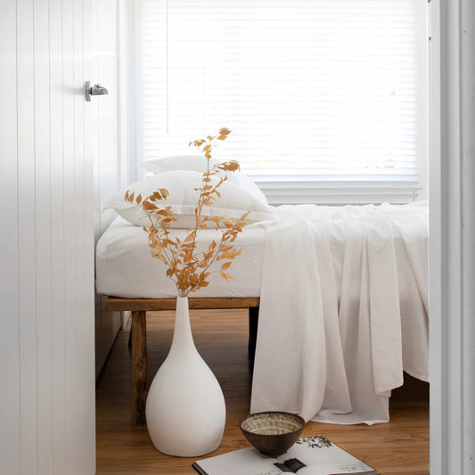 White vase with gold leaves on a wooden stool in a room with white walls and a window.