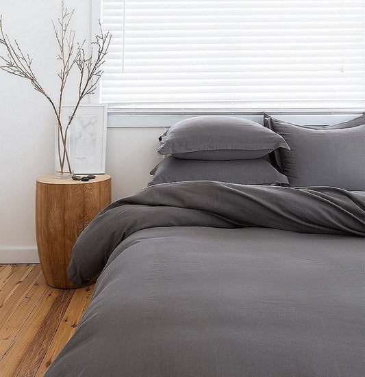 Gray bedding set on a bed with a wooden side table and plant in the background.