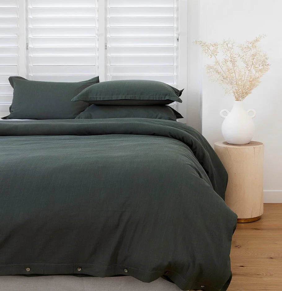 Dark green bedding set on a bed with a side table and decorative vase in the background.