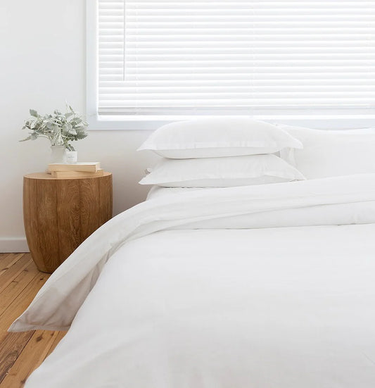 White bedding on a bed in a bedroom with a wooden side table and plant.