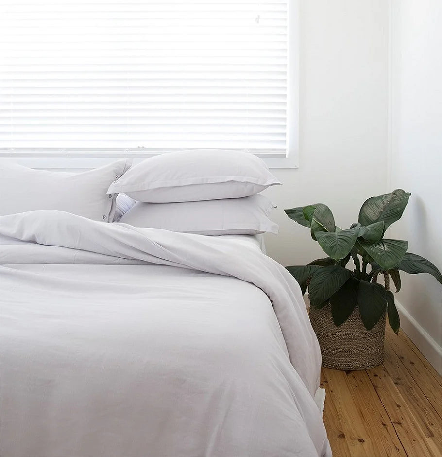 Bed with white bedding and pillows in a room with a plant and wooden floor.
