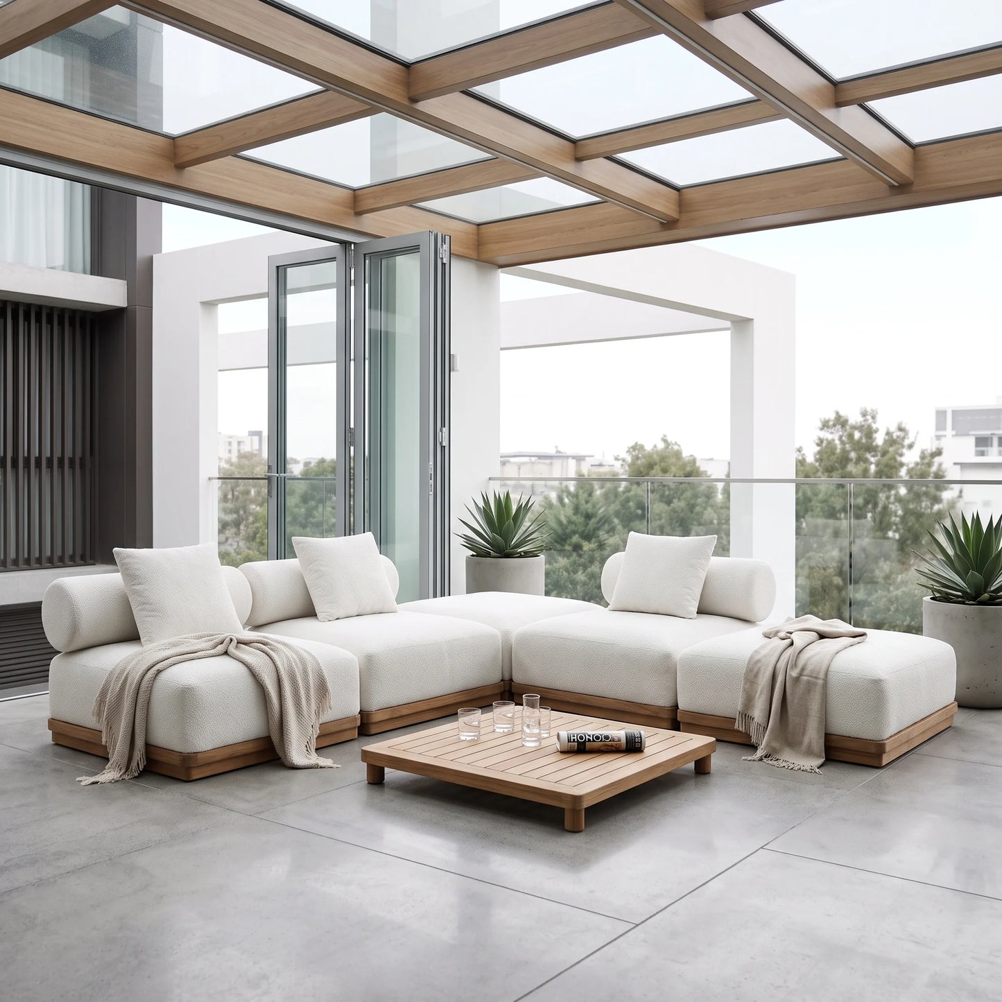 A modern outdoor sectional sofa in off-white fabric with light wood frames, arranged on a spacious concrete patio under a glass-roofed pergola. A matching wooden coffee table sits in front, with neutral throw blankets draped over the sofa cushions.