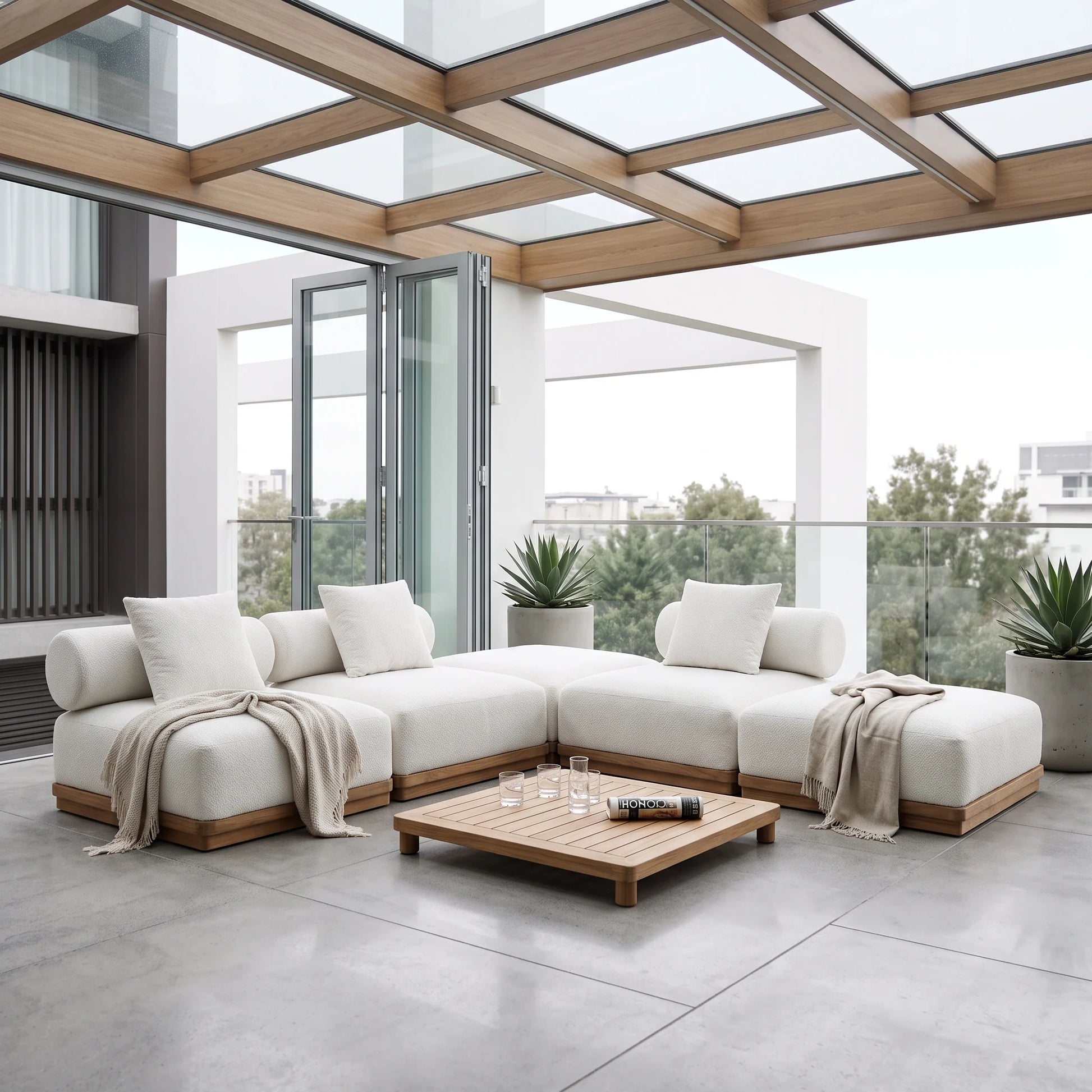 A modern outdoor sectional sofa in off-white fabric with light wood frames, arranged on a spacious concrete patio under a glass-roofed pergola. A matching wooden coffee table sits in front, with neutral throw blankets draped over the sofa cushions.