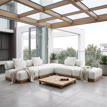 A modern outdoor sectional sofa in off-white fabric with light wood frames, arranged on a spacious concrete patio under a glass-roofed pergola. A matching wooden coffee table sits in front, with neutral throw blankets draped over the sofa cushions.