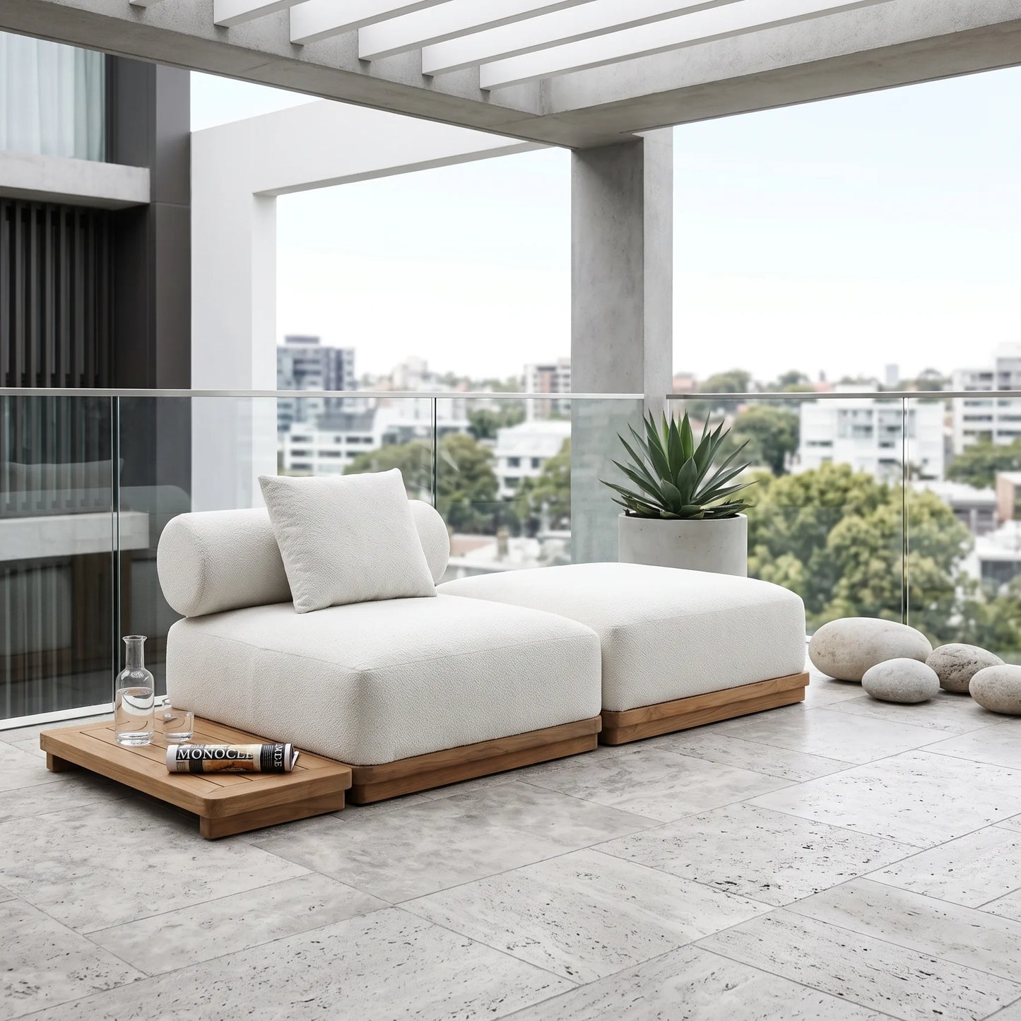 A modern outdoor sectional sofa in off-white fabric with a light wood base, styled on a stone-tiled balcony with a matching wooden tray, a potted plant, and decorative stones, overlooking a city skyline.