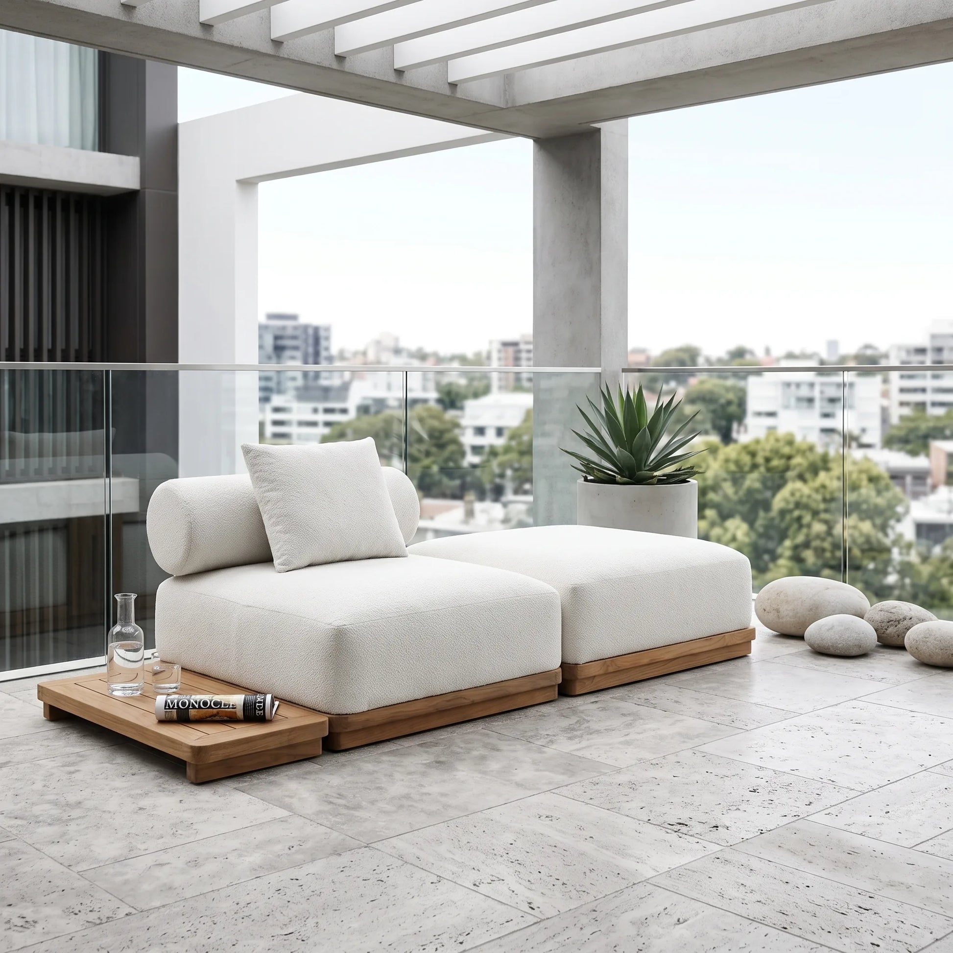 A modern outdoor sectional sofa in off-white fabric with a light wood base, styled on a stone-tiled balcony with a matching wooden tray, a potted plant, and decorative stones, overlooking a city skyline.