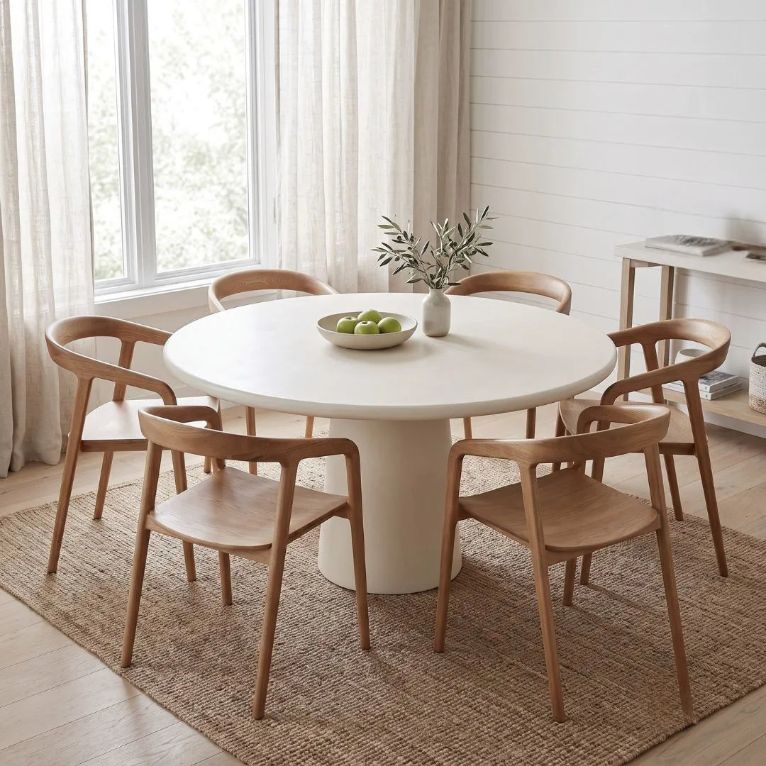 A round white dining table with a pedestal base surrounded by six light wood chairs, set on a woven rug in a bright, modern dining room.