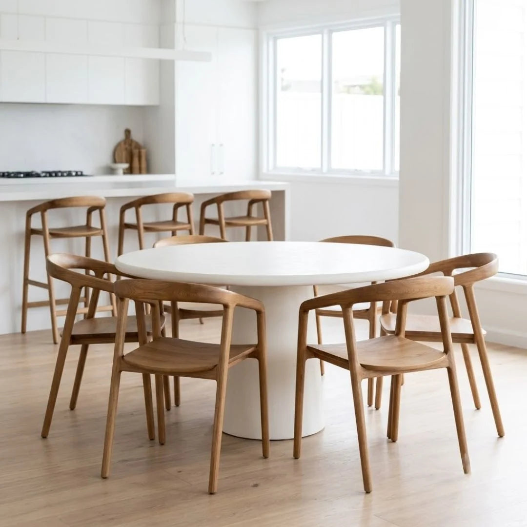 A round white dining table with a white pedestal base surrounded by six wooden chairs with curved backrests, set in a bright, modern kitchen with white cabinets and large windows.