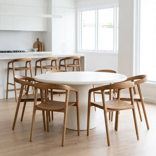 A round white dining table with a white pedestal base surrounded by six wooden chairs with curved backrests, set in a bright, modern kitchen with white cabinets and large windows.