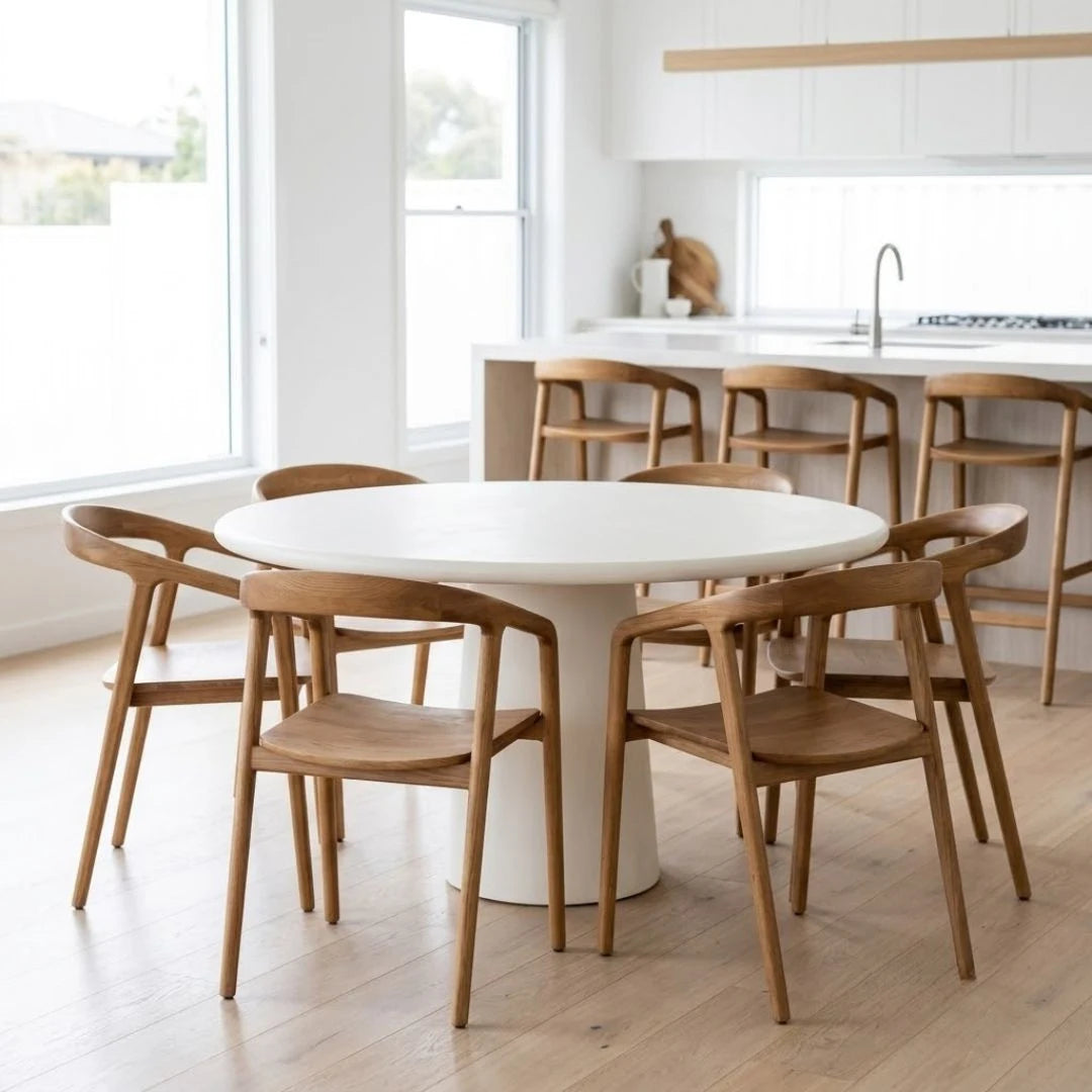 A round white dining table with a white pedestal base surrounded by six wooden chairs with curved armrests, set in a bright, modern kitchen with light wood floors and white cabinetry.
