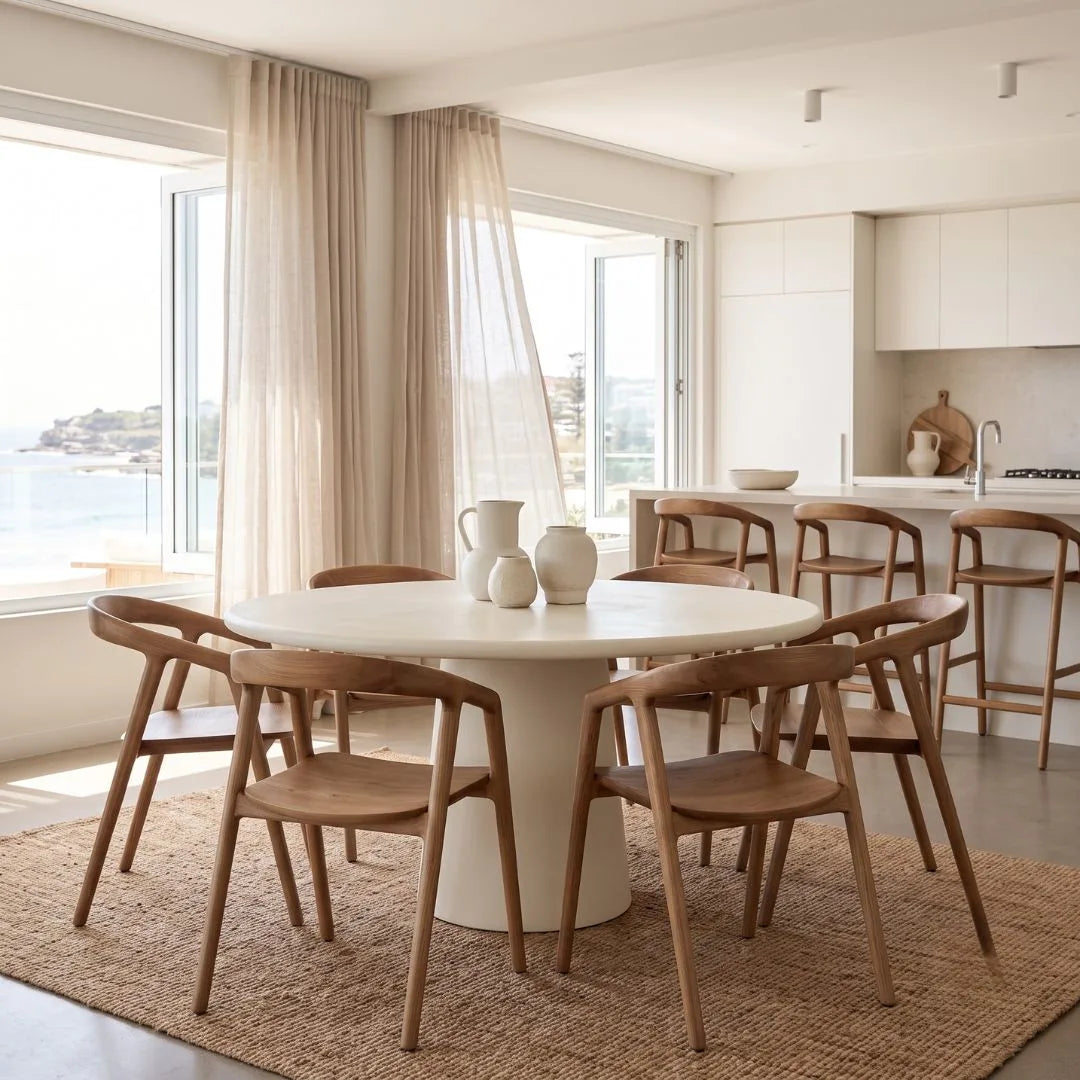 A modern dining room setup featuring a round white pedestal table surrounded by six wooden chairs with curved backs, set on a woven jute rug. The room has large windows with sheer curtains, a view of the sea, and a contemporary kitchen in the background.