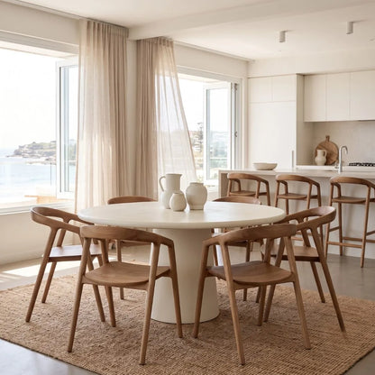 A modern dining room setup featuring a round white pedestal table surrounded by six wooden chairs with curved backs, set on a woven jute rug. The room has large windows with sheer curtains, a view of the sea, and a contemporary kitchen in the background.