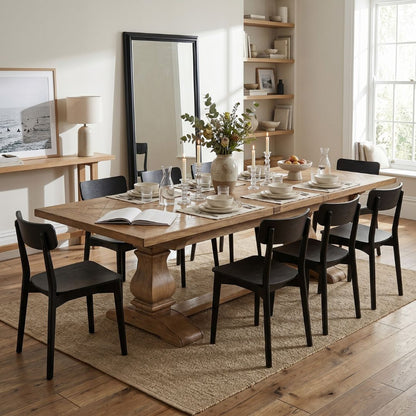 A rectangular wooden dining table with a turned pedestal base and trestle-style feet, surrounded by black chairs in a bright, modern dining room. The table is set with white plates, glassware, and a vase of greenery, placed on a woven jute rug over hardwood floors.