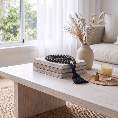 A styled interior scene showing a light wood coffee table with stacked books, a black beaded tassel necklace draped over them, a beige ceramic vase with dried grass stems, and a candle on a woven placemat near a window and sofa.