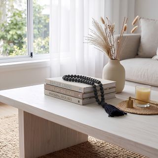 A light wood coffee table in a bright living room setting, styled with stacked books, a black beaded tassel necklace, a beige ceramic vase with dried grasses, and a lit candle on a woven coaster.