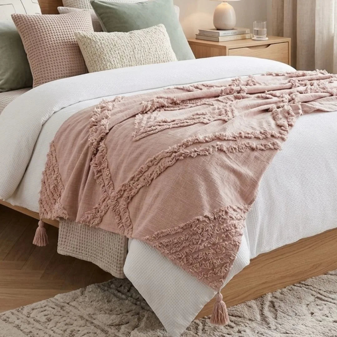 Blush Pink textured blanket draped over a bed with pillows and a nightstand in the background.