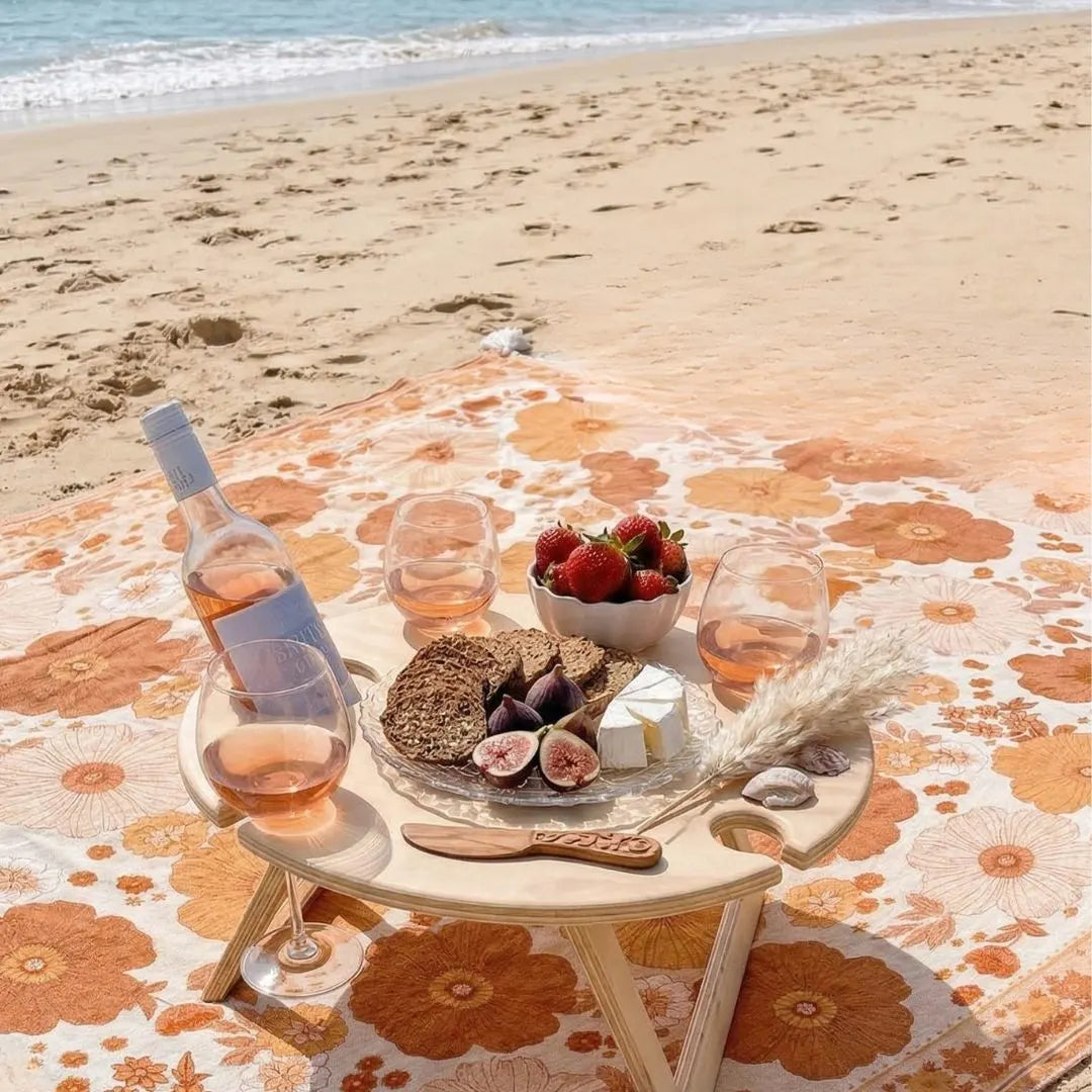 Beach picnic setup with a bottle of rosé, glasses, fruits, and bread on a floral blanket.