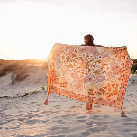 Person holding a large floral-patterned towel on a sandy beach at sunset.