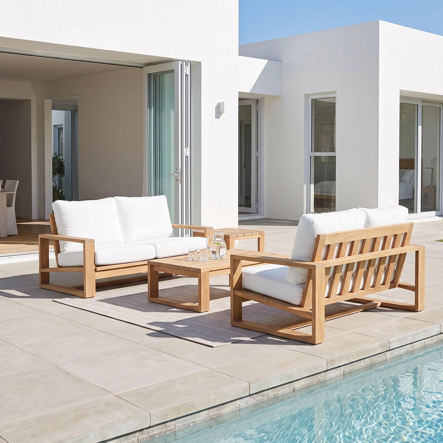 A modern outdoor patio furniture set with a wooden three-seat sofa, matching armchair, and low rectangular coffee table, all with thick white cushions, arranged on a light stone tile deck next to a swimming pool.
