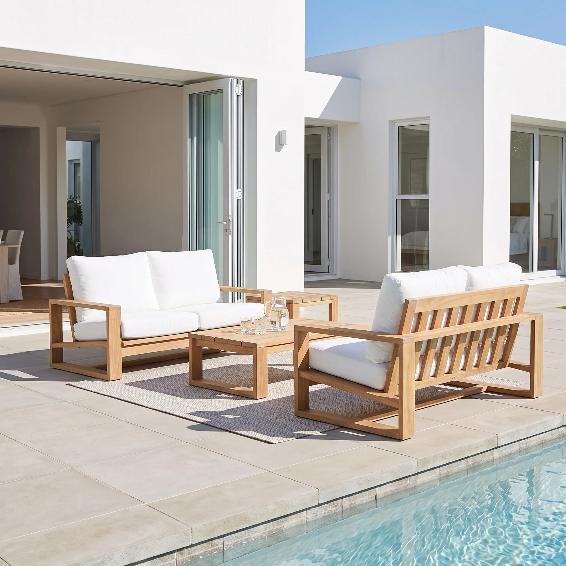 A modern outdoor patio furniture set with a wooden three-seat sofa, matching armchair, and low rectangular coffee table, all with thick white cushions, arranged on a light stone tile deck next to a swimming pool.