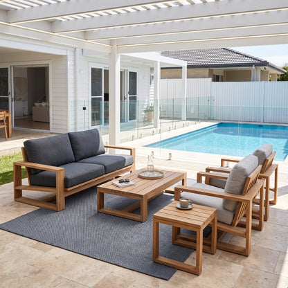 Outdoor patio furniture set including a wooden loveseat, armchair, coffee table, and side table with gray cushions, arranged on a rug beside a swimming pool under a white pergola.