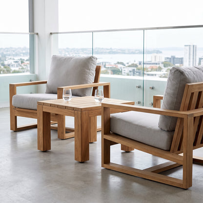 outdoor patio furniture set featuring two wooden armchairs with light gray cushions and a matching wooden side table, arranged on a concrete balcony with a city and water view.