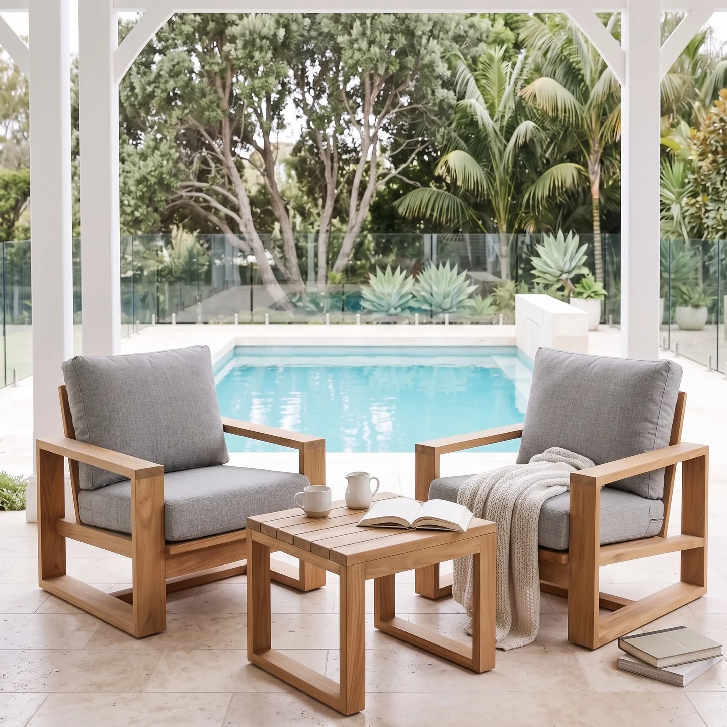 Two wooden outdoor armchairs with gray cushions and a matching wooden side table, arranged under a covered patio overlooking a swimming pool and tropical garden.