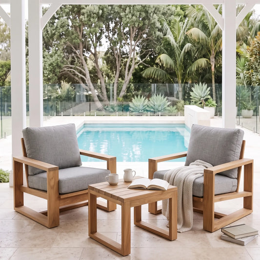 Two wooden outdoor armchairs with gray cushions and a matching wooden side table, arranged under a covered patio overlooking a swimming pool and tropical garden.
